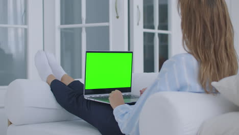 A-young-woman-sits-with-a-laptop-on-her-lap-with-a-green-screen-during-quarantine.-chromakey-on-the-laptop-screen.-Make-a-video-conference-and-talk-to-the-green-screen